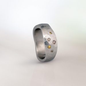Platinum hammered band with an organic, sculptural shape, set with scattered coloured diamonds in varying sizes.