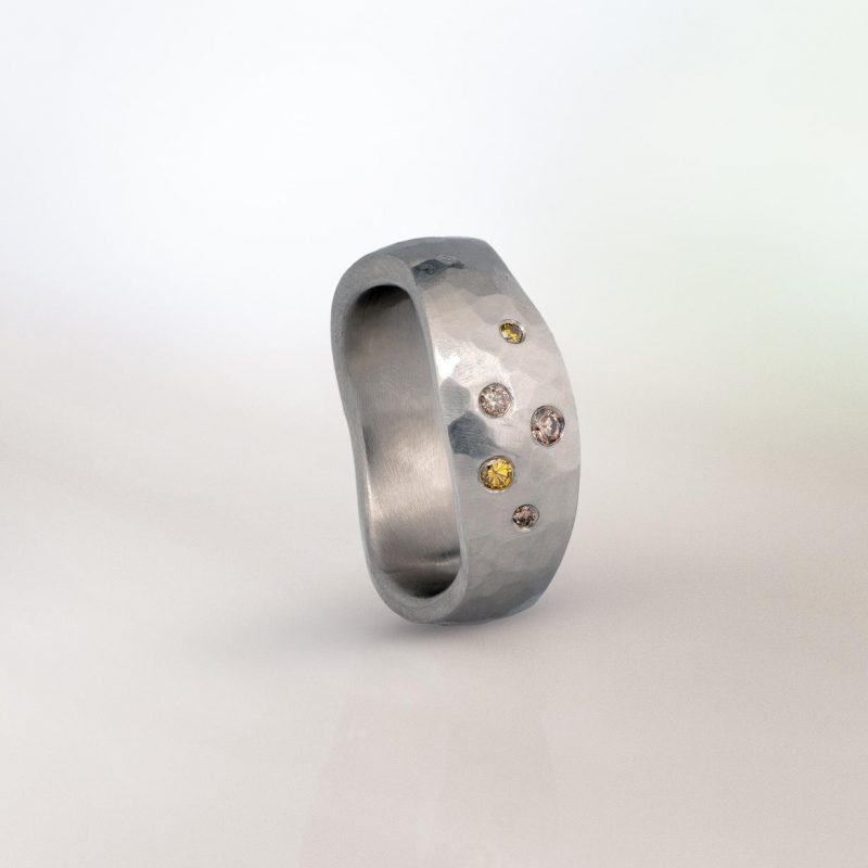 Platinum hammered band with an organic, sculptural shape, set with scattered coloured diamonds in varying sizes.