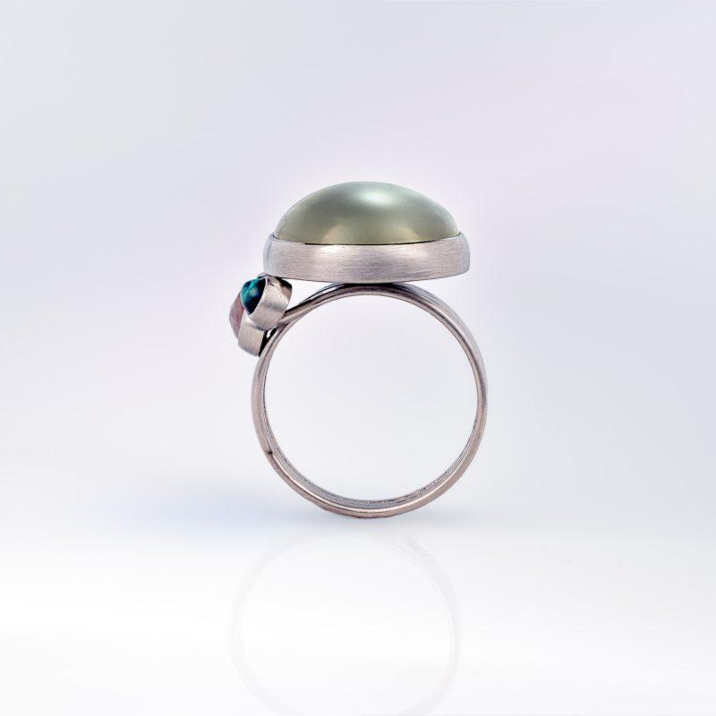 Underside ide view of a platinum Ribbon ring with a wide, tiered band, showing the domed cabochon moonstone rising above the setting, with an orange pear-shaped rose-cut diamond and a green cabochon tourmaline shown off-set to the left side of the moonstone.