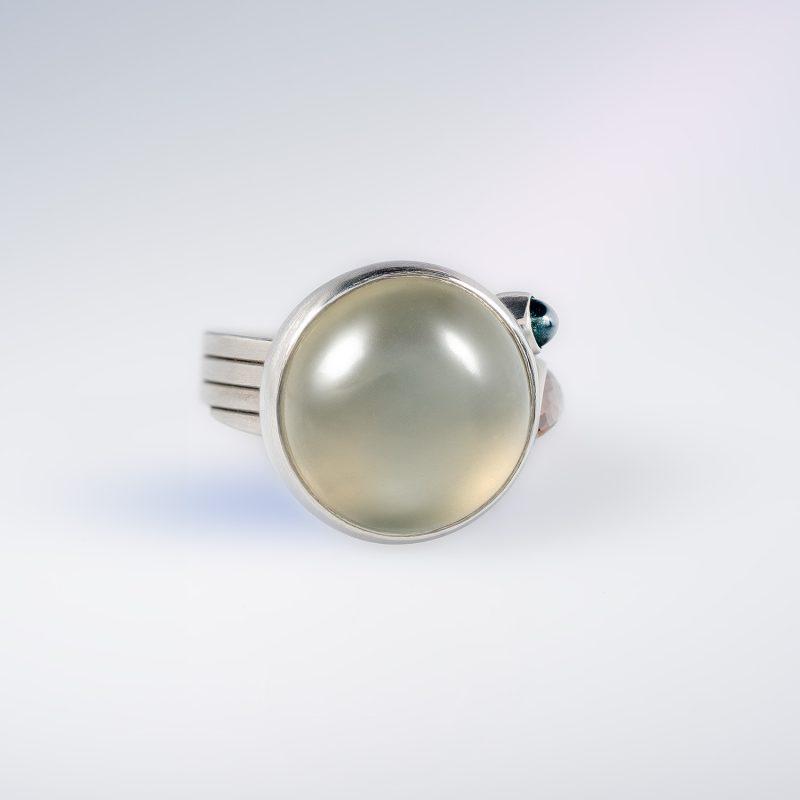 Platinum ring with a wide, ribbon-wrapped band, featuring a large cabochon moonstone, an orange pear-shaped rose-cut diamond, and a small green cabochon tourmaline.