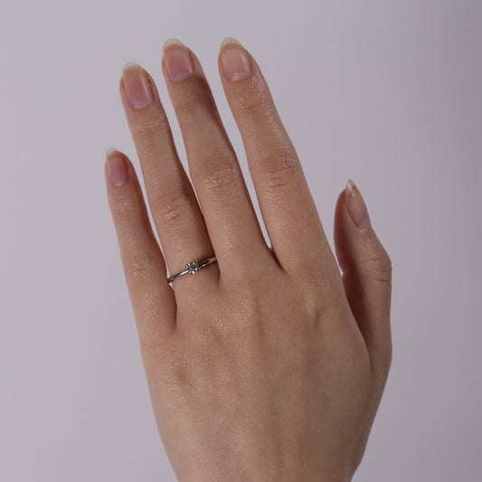 A close-up of a person's hand wearing a delicate silver ring with a small clear gemstone on the ring finger, set against a plain light gray background.